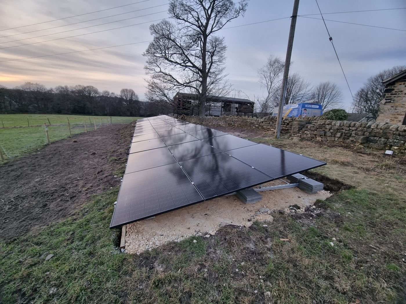 A low row of black solar panels on a gravel base next to a field and stone wall