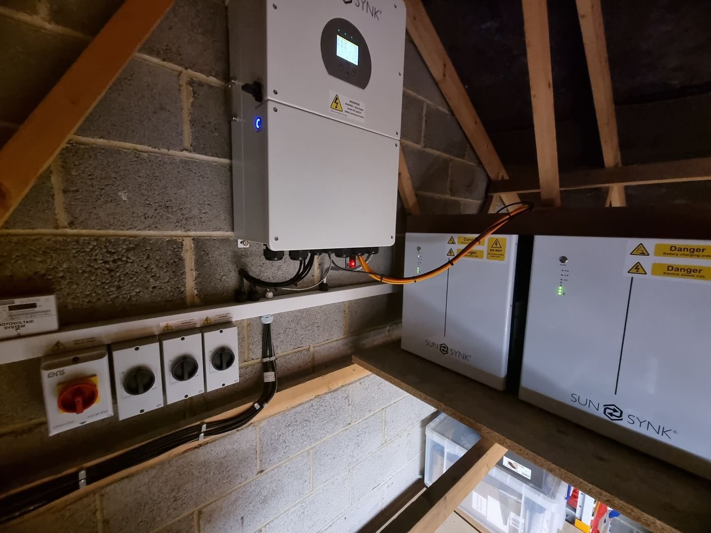 A Sunsynk hybrid inverter with two Sunsynk batteries and isolator switches in a loft space